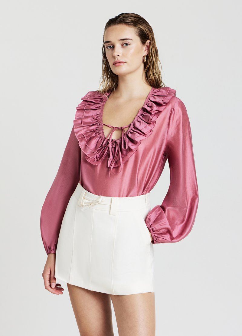 Blusa di raso rosa con scollatura ruffle e dettaglio con fiocco, abbinata a una mini gonna bianca a vita alta con cintura e cuciture strutturate.