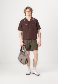 Braunes Kurzarmhemd mit weißen Akzenten, grüne Shorts, graue Tasche mit mehreren Taschen, weiße Sneaker und weiße Socken.