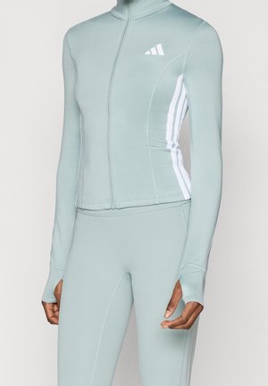 Femme portant une veste ajustée bleu clair avec fermeture éclair et des leggings avec des trous pour le pouce et des bandes blanches sur les côtés de la veste.