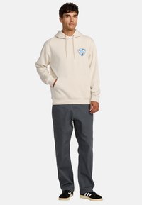 Crème hoodie met een voorzak en een blauw logo, gecombineerd met donkergrijze broek en zwarte schoenen met witte strepen. Zachte stoftextuur.