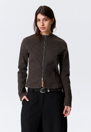 Γυναίκα με καστανά μαλλιά φορεί σκούρο καφέ μπουφάν με φερμουάρ και μαύρο παντελόνι, όρθια με το ένα χέρι στην τσέπη, μπροστά σε απλό φόντο.