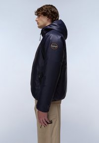 Marineblauwe pufferjack met een capuchon, glanzende textuur, ritszakken en een ronde logo patch op de linkermouw. Lange mouwen en een comfortabele pasvorm.