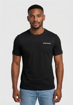 Uomo con indosso una maglietta nera Sergio Tacchini e jeans azzurri, in piedi davanti a uno sfondo grigio chiaro uniforme.