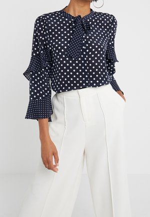 Kobieta w granatowej bluzce w kropki z zawiązaną kokardą i falbanami na rękawach, w połączeniu z wysokim stanem i szerokimi białymi spodniami.