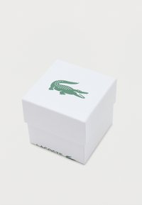 Bílý čtvercový box s texturovaným povrchem, se zeleným logem krokodýla a textem "LACOSTE" na boku. Uzavřený design.