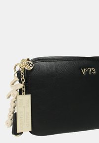 La borsa a tracolla nera in finta pelle presenta una chiusura con zip, hardware dorato e etichette decorative con testo. Forma rettangolare compatta.