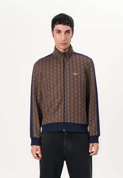 Veste zippée marron à motifs avec des accents marins, col haut, poignets côtelés et un petit logo. Le tissu a une texture lisse.