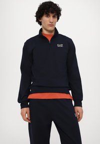 Tumed sinine tõmblukuotsa sweatshirt, mis on valmistatud puuvilla segust, pikad varrukad, soonikkoes mansetid ja rinnal EA7 logo.