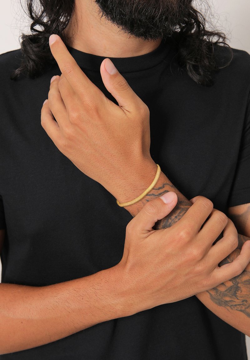 Gold-colored bracelet made of twisted wire, worn on a wrist with a tattoo, hands positioned with one finger pointed upward.