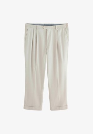 Fiatal férfi göndör hajjal, krémszínű kétsoros öltönyben, kék ingben és fekete cipőben, egy mintás szőnyegen áll, háttérben egy faburkolatos fallal.