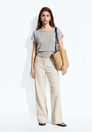 Mujer joven con cabello largo y castaño que lleva una camiseta gris de manga corta, pantalones anchos beige, sandalias negras y lleva un bolso tote tejido.