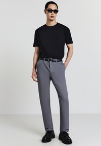 Uomo che indossa occhiali da sole neri, t-shirt nera, pantaloni grigi con cintura nera e scarpe nere, in piedi con una mano in tasca.