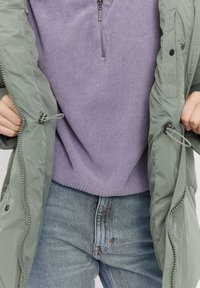 Grøn polstret jakke med snore, parret med en tekstureret lilla sweater og lyseblå denimjeans med subtile slidmærker.