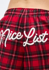 Κόκκινο παντελόνι πιτζάμας σε καρό σχέδιο με λευκό κείμενο "Νice List" στην πίσω πλευρά και ένα μικρό γραφικό φιόγκου κοντά στο κείμενο.