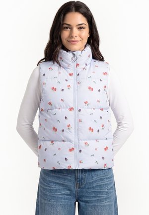 Giovane donna che indossa un gilet imbottito azzurro chiaro con motivo floreale e cursore a forma di cuore sopra una camicia bianca a maniche lunghe e jeans blu.