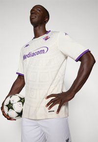 Weißes Fußballtrikot mit subtiler Musterstruktur, lila Akzenten an den Ärmeln und am Ausschnitt, mit dem Logo "Mediacom" versehen, kombiniert mit weißen Shorts und einem mehrfarbigen Ball.