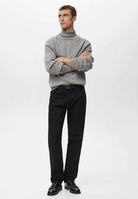 Grijze geribbelde coltrui, zwarte broek met rechte pijpen en gepoetste zwarte schoenen. Model tegen een neutrale achtergrond, met gekruiste armen.