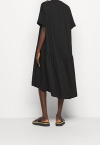 Person wearing loose, black, knee-length dress with gathers near hem and black flat sandals, standing against a plain white background.