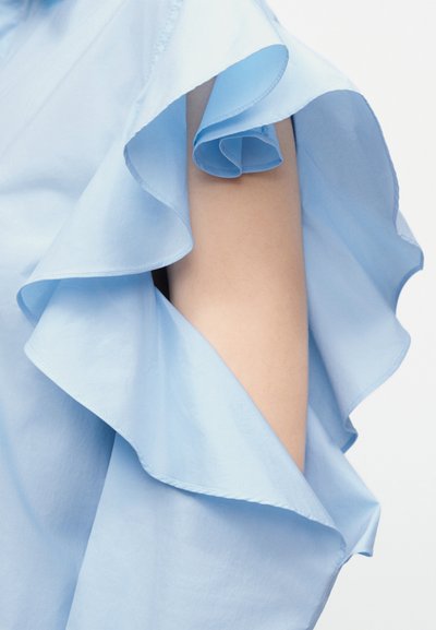 Tissu bleu clair avec des volants superposés drapés sur un bras, sur fond blanc, mettant en valeur la texture et les plis doux.