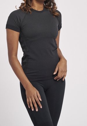 Vrouw die een nauwsluitend zwart T-shirt met korte mouwen en zwarte legging draagt, staand met één hand op haar heup en de andere langs haar dij.