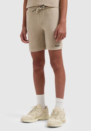 Persoon draagt beige badstofshorts met trekkoord, witte sportsokken en beige sneakers, staand tegen een effen achtergrond.
