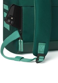 Groene stoffen tas met een gestructureerde afwerking, voorzien van een ritsvak waarin een zwarte smartphone is opgeborgen, en een brede verstelbare schouderband met hardware.