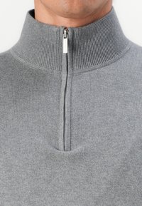 Sivi zip-up dukserica s visokim rebrastim ovratnikom, sa zatvaračem na pola dužine i glatkom teksturom. Oznaka brenda vidljiva na povlačenju zatvarača.