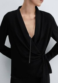 Top avvolgente nero con scollatura a V profonda e maniche lunghe, dotato di un laccio in vita e una lunga collana con pendente in argento.