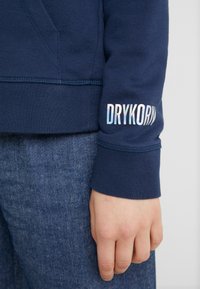 Felpa blu navy con polsini a coste, caratterizzata da un logo iridescente con la scritta "DRYKORN" sulla manica. Jeans in denim visibili sotto.