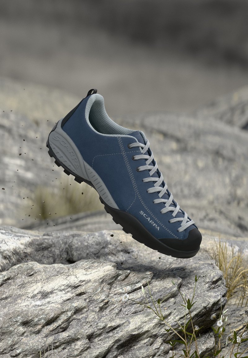 Μπλε και γκρι παπούτσι πεζοπορίας με κορδόνια που αιωρείται πάνω από βραχώδες έδαφος, παρουσιάζοντας την πέλμα και τον ανθεκτικό σχεδιασμό για εξωτερικούς χώρους.