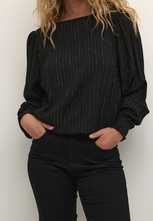 Femme portant un chemisier noir à manches longues avec de fines rayures verticales argentées et un pantalon noir, les mains dans les poches.