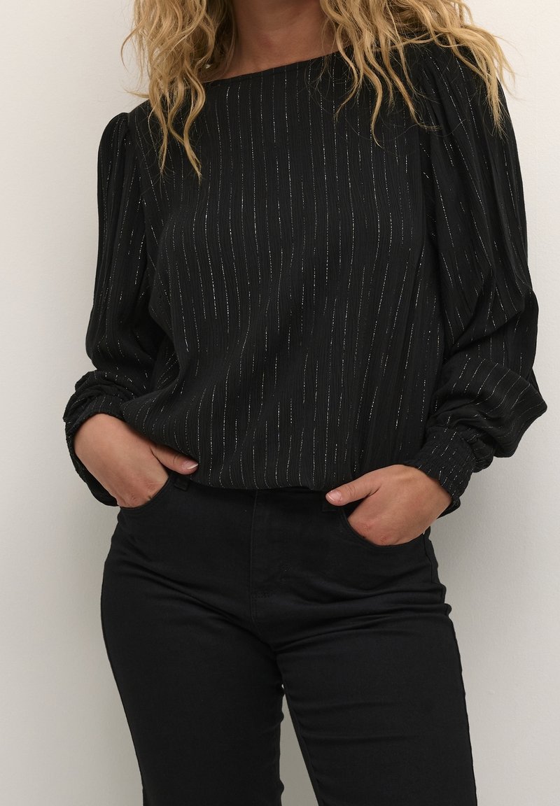 Femme portant un chemisier noir à manches longues avec de fines rayures verticales argentées et un pantalon noir, les mains dans les poches.