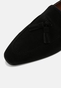 Mocassin en daim noir avec un bout carré arrondi et deux pampilles en daim sur le dessus, couture détaillée autour du bord avant.