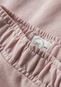 Lichtroze stof heeft geplooide details en een zichtbare plastic knop, met een label dat gedeeltelijk zichtbaar is bij de naad.