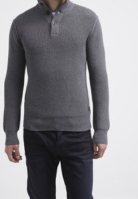 Uomo che indossa un maglione grigio lavorato a maglia con colletto abbottonato e pantaloni scuri, in piedi davanti a uno sfondo bianco semplice.