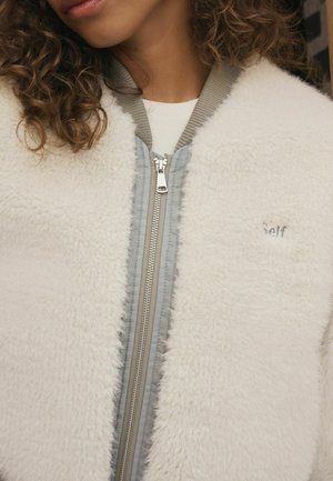 Valkoinen fleece-bomber-takki, jossa on harmaa vetoketju edessä, röyhelökaula ja huomaamaton "Self"-kirjonta rinnassa. Pehmeä tekstuuri koko takissa.