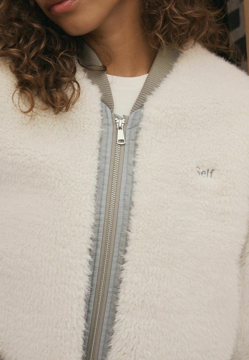 Biała bomberka z polaru z szarym zamkiem z przodu, prążkowanym kołnierzem i subtelnym haftem "Self" na piersi. Miękka faktura na całej powierzchni.