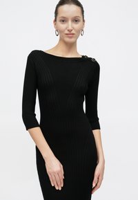 Γυναίκα που φοράει εφαρμοστό μαύρο πλεκτό φόρεμα με μανίκια μέχρι τον αγκώνα και δύο κουμπιά στον αριστερό ώμο, στέκεται μπροστά σε λευκό φόντο.