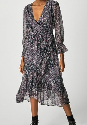 Femme portant une robe portefeuille fleurie longueur genoux avec un ourlet volanté et des manches longues transparentes, associée à des bottines noires et des chaussettes.