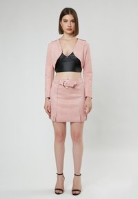 Rosa Wildleder-Cropped-Jacke kombiniert mit einem schwarzen Cropped-Top und einem rosa Wildleder-Minirock mit einem taillierten Bund und fransigen Kanten. Schwarze Knöchelriemen.