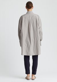 Lange, hellgraue Hemdjacke aus strukturiertem Stoff, mit einem Rückenfalte, geknöpften Manschetten und einem klassischen Kragenstil. Kombiniert mit dunklen Hosen.
