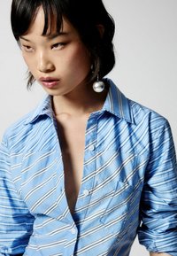 Cămașă bleu deschis, cu dungi și guler, având un buzunar pe piept; prezintă dungi albe și negre; fabricată dintr-un bumbac neted; desfăcută la guler.