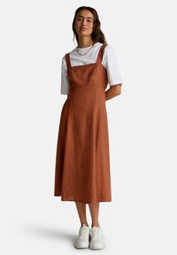 Mujer con un vestido midi sin mangas color óxido sobre una camiseta blanca de mangas cortas, zapatillas blancas y un collar de cadena dorada, de pie con las manos detrás de la espalda.