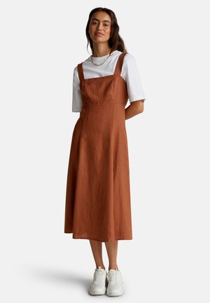 Donna che indossa un abito midi senza maniche color ruggine sopra una camicetta bianca a maniche corte, sneakers bianche e collana d'oro, in piedi con le mani dietro la schiena.