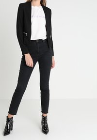 Musta räätälöity bleiseri vetoketjullisilla taskuilla, yhdistetty valkoiseen t-paitaan, jossa on violettia tekstiä. Mustat korkeavyötäröiset farkut ja kiiltävät nilkkurit.