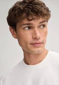 Biała bluza z okrągłym dekoltem, o teksturowanym wykończeniu, wykonana z lekkiego materiału, z ribbowanym kołnierzykiem, noszona przez modela z krótkimi, falującymi brązowymi włosami.