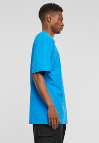 Zila kokvilnas T-kreklu ar īsām piedurknēm, apaļu kakla izgriezumu un brīvu piegriezumu, attēlots uz personas sānskata fona uz neitrāla fona.