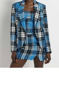 Žena v modré, černé a bílé kostkované saku přes odpovídající bezrukávový top a asymetrickou mini sukni.