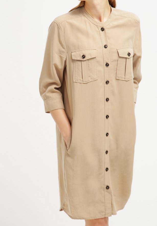 Robe beige boutonnée avec ourlet arrivant aux genoux, deux poches avant à rabat boutonné et manches trois-quarts, portée par une personne les mains dans les poches latérales.
