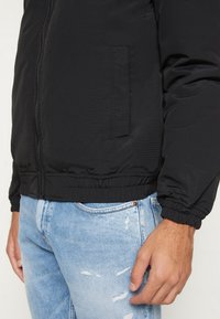 Giacca bomber nera con zip, polsini e vita elasticizzati; indossata sopra jeans chiari strappati. Presenta una tasca laterale.